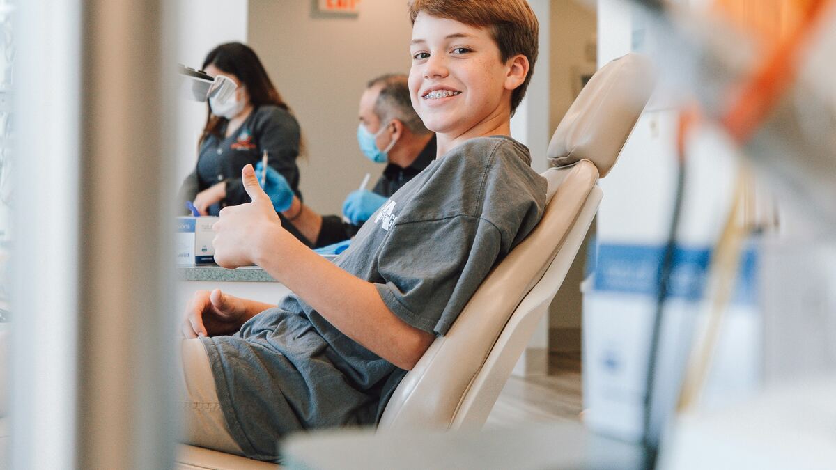 A boy in a dental chair grinning and giving a thumbs up